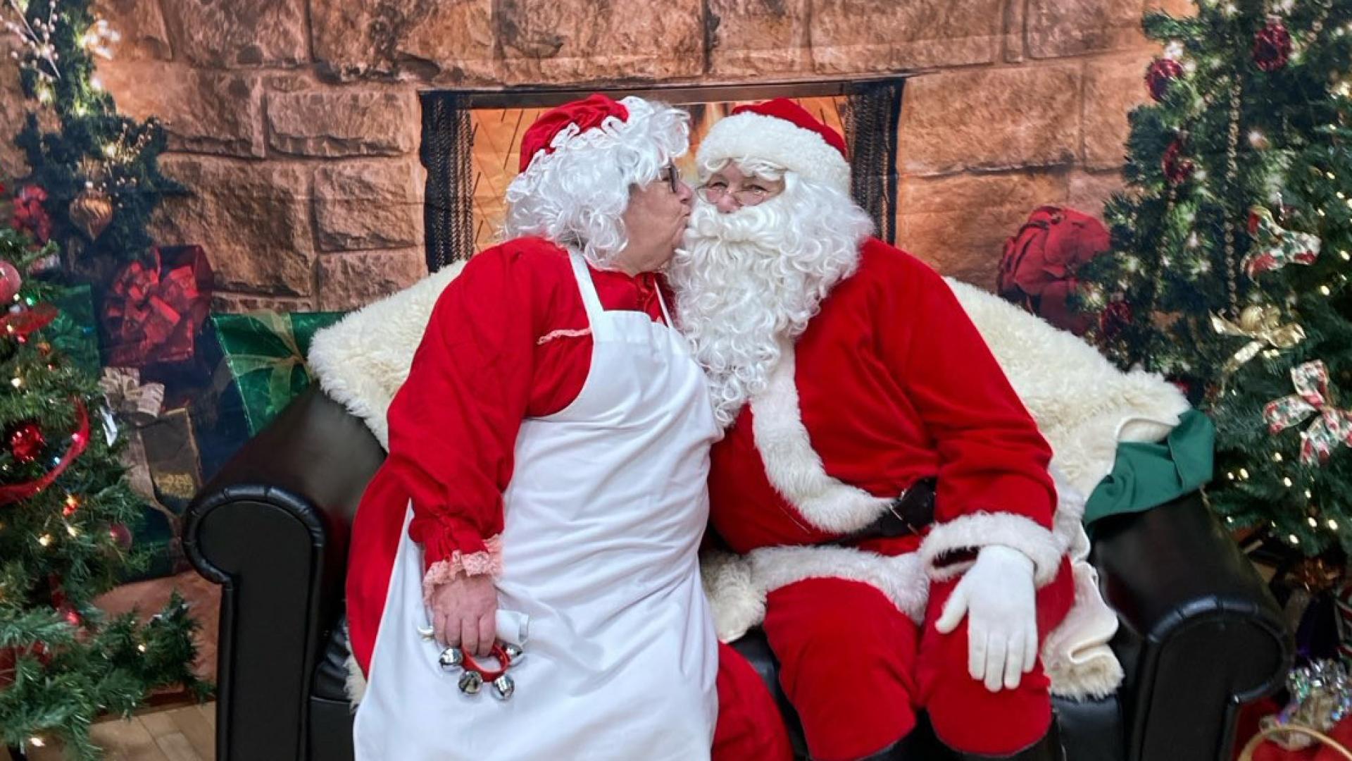 Mrs. Claus sits on Santa’s lap, giving kiss on the cheek as they smile warmly in front of a glowing fireplace.