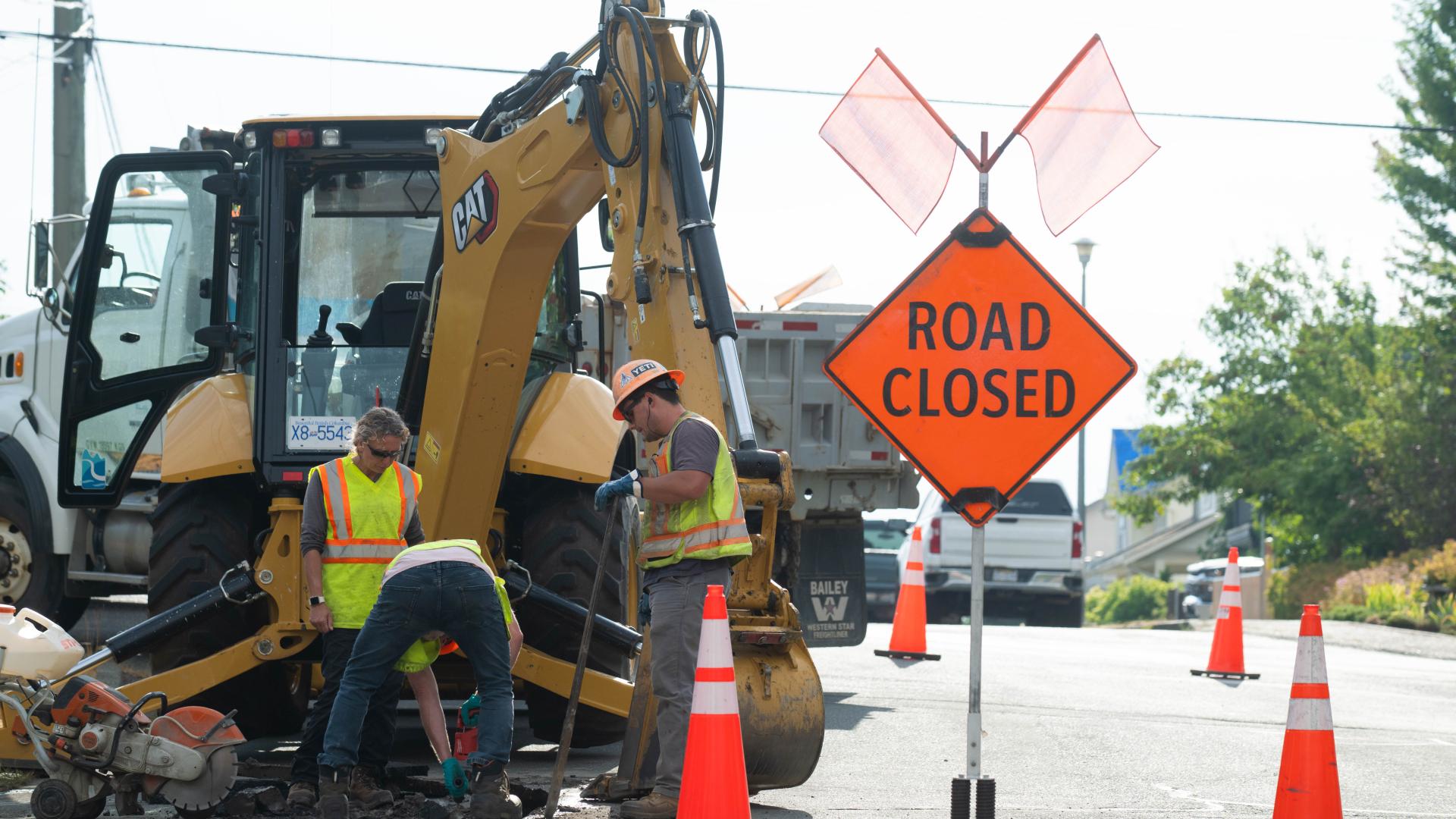 Construction workers and large truck digging hole in road with a sign that reads Road Closed and pylons surrounding worksite.