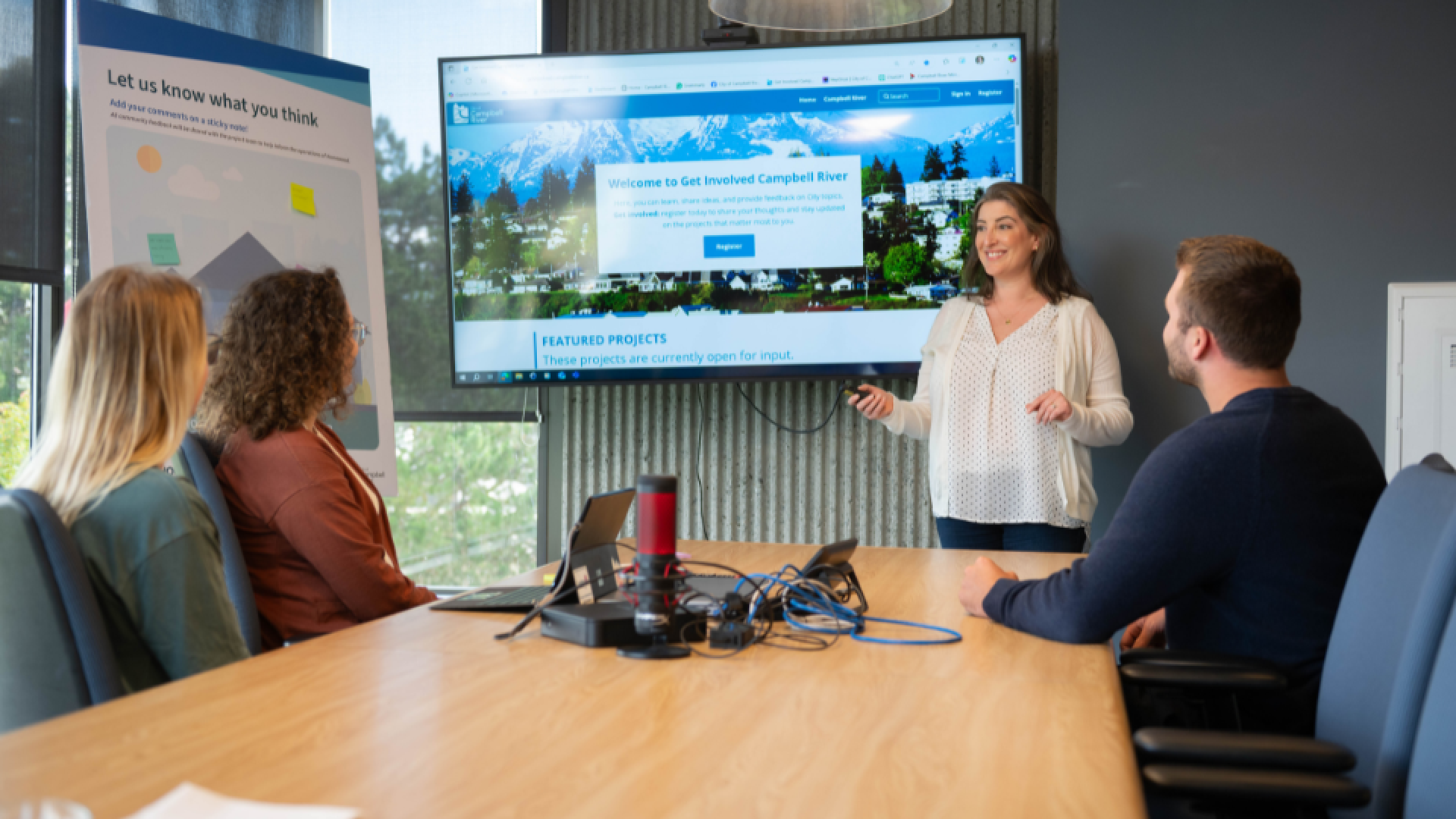 Person standing and presenting in front of a large screen with three people sitting at a table.