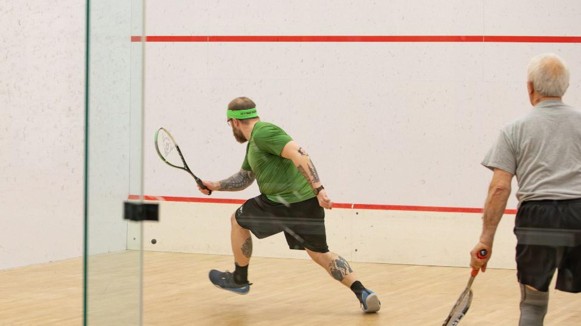 Two squash players competing on an indoor court, both holding racquets, with the ball in mid-air near the front wall