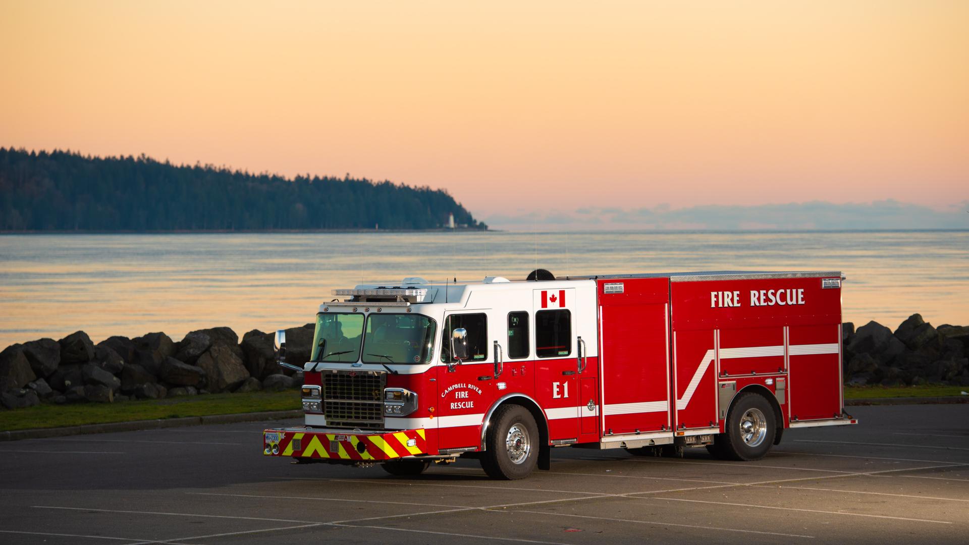 A red fire engine is parked in a parking lot during sunset with the ocean and an island in the background.