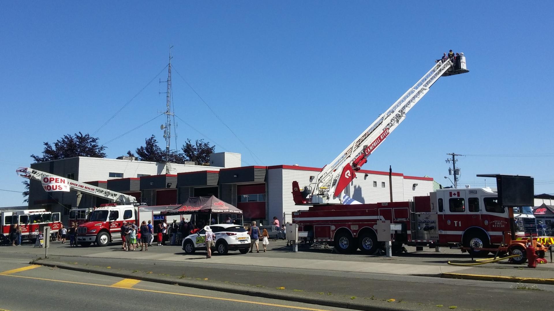 A fire station with fire engines and ladder trucks parked out front during a sunny day with blue skies. The ladders are raised into the sky, and an Open House sign is displayed. Members of the public are walking around.