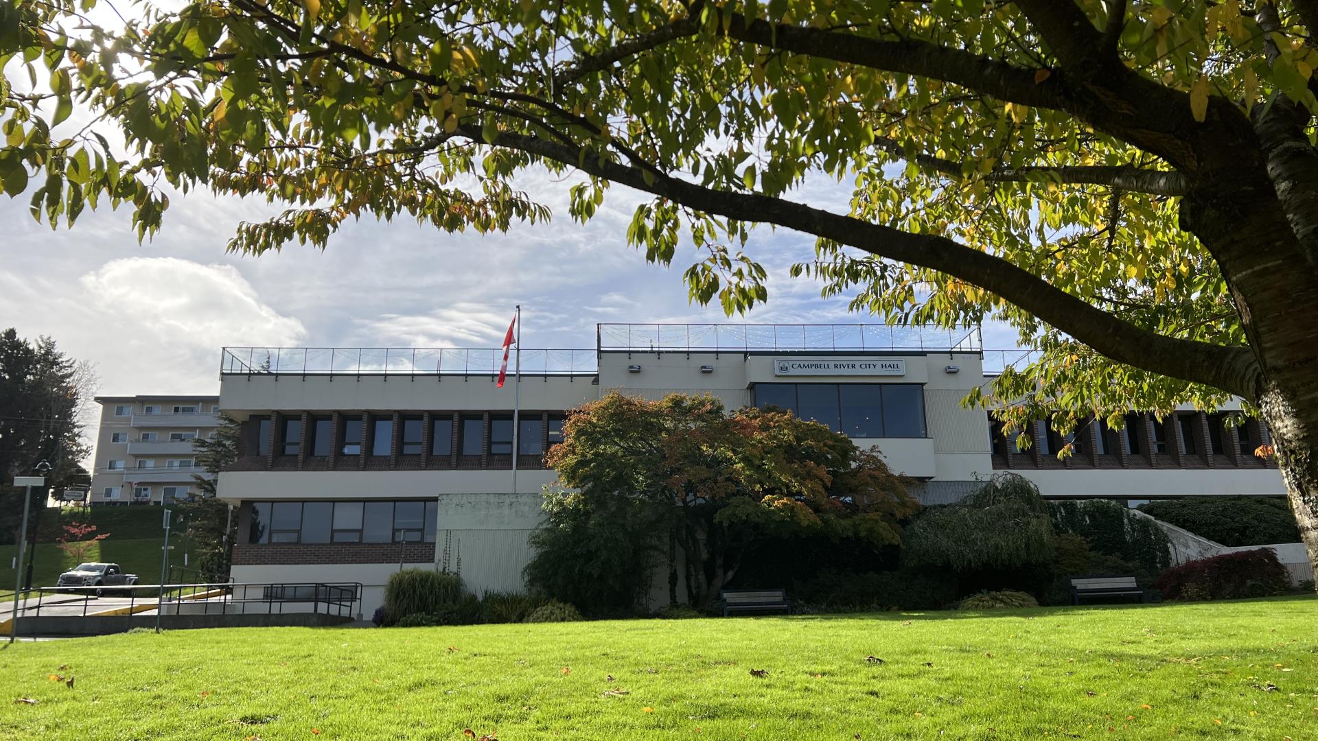 Exterior view of Campbell River's City Hall building. A Canada flag flies in front of the building. Greenery, a lawn and a leafy tree are in the foreground.