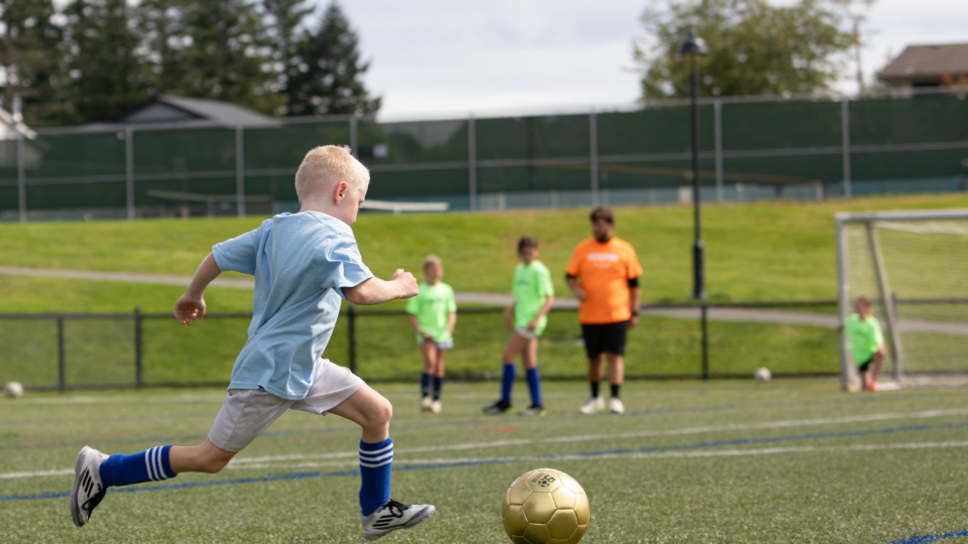 A child kicks a soccer ball on a field while other players are visible in the background.