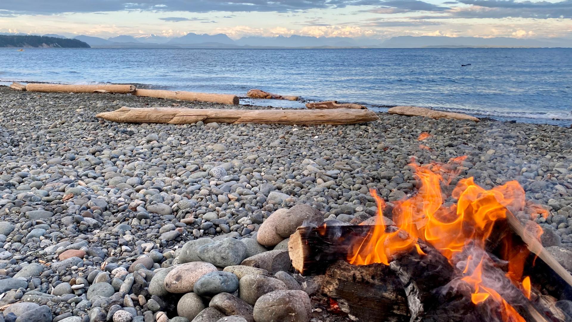 A beach fire during sunset with driftwood, the ocean and scattered clouds in the background.