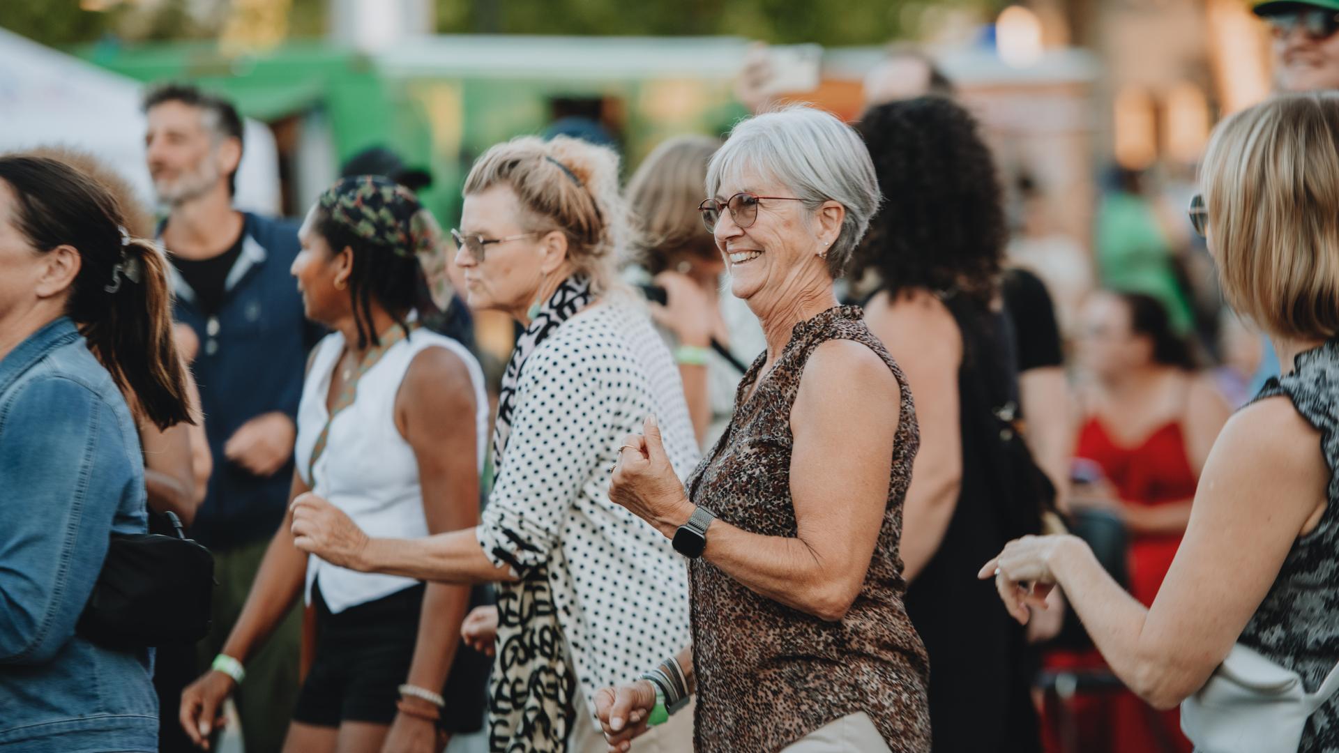 A crowd dances at an outdoor concert on a sunny day. They are smiling and laughing, and the atmosphere is festive.