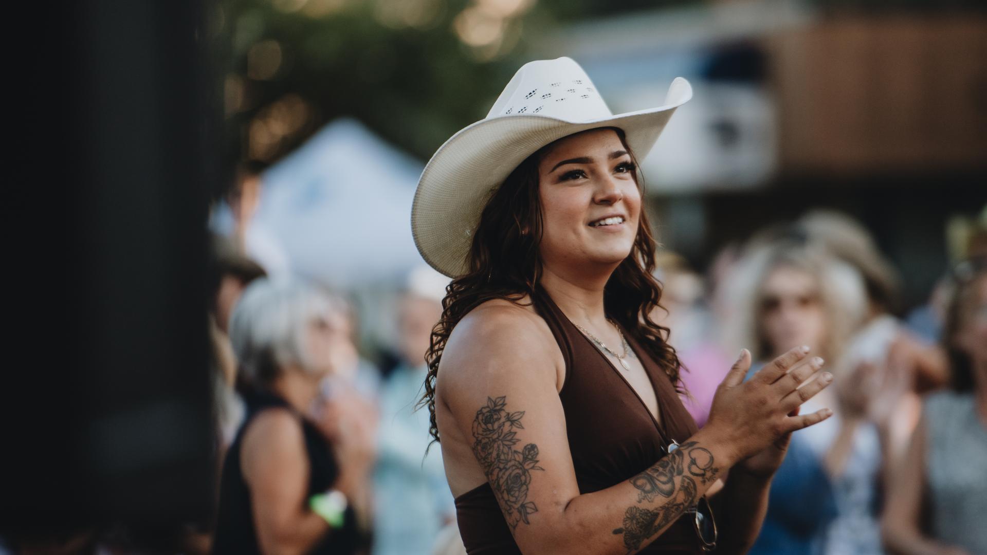 The foreground features a person wearing a cowboy hat who is clapping along to music at an outdoor concert. A crowd dances in the background.