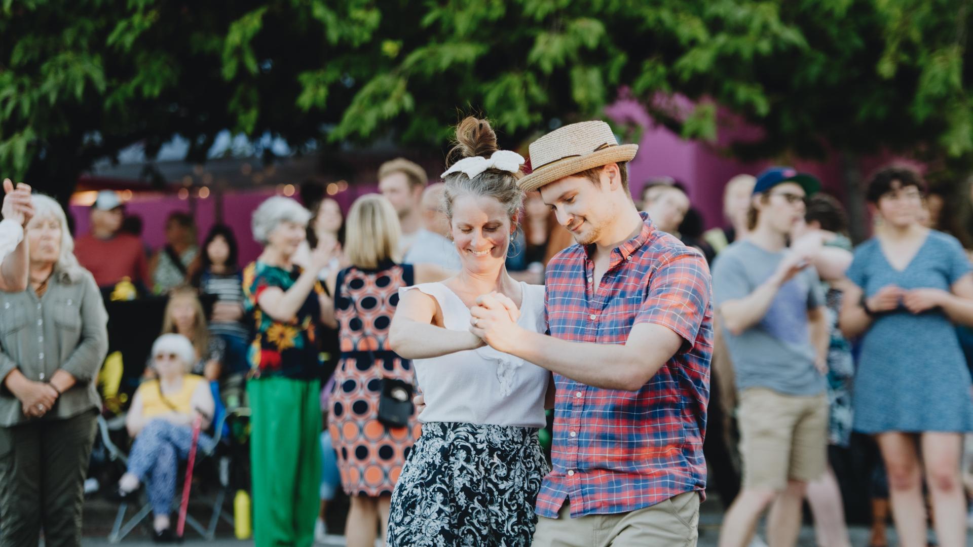 A couple dance outdoors at a concert, surrounded by a crowd. A building and trees are in the background.