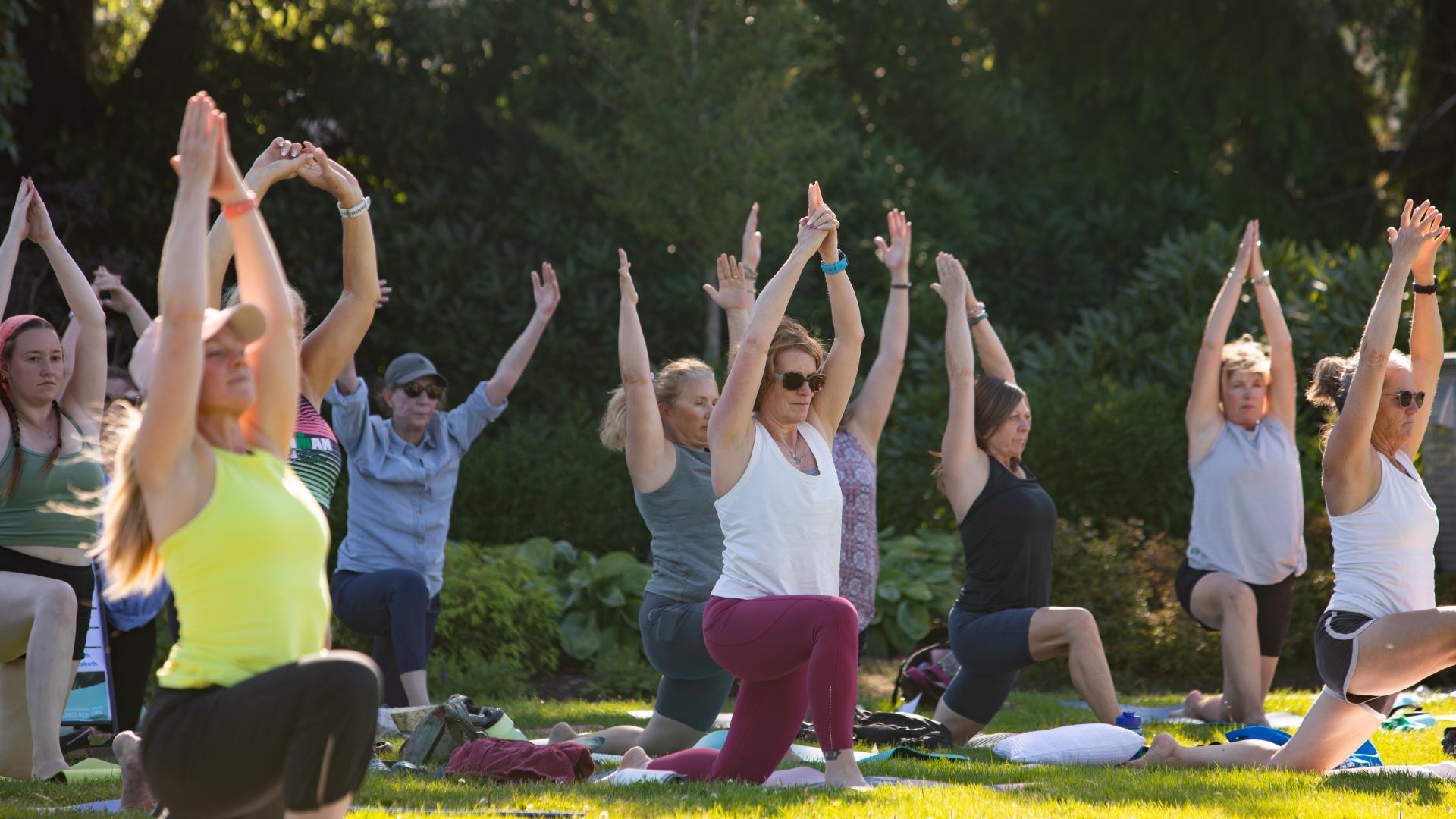 People doing yoga posses on mats on the grass in a field with trees in the background.