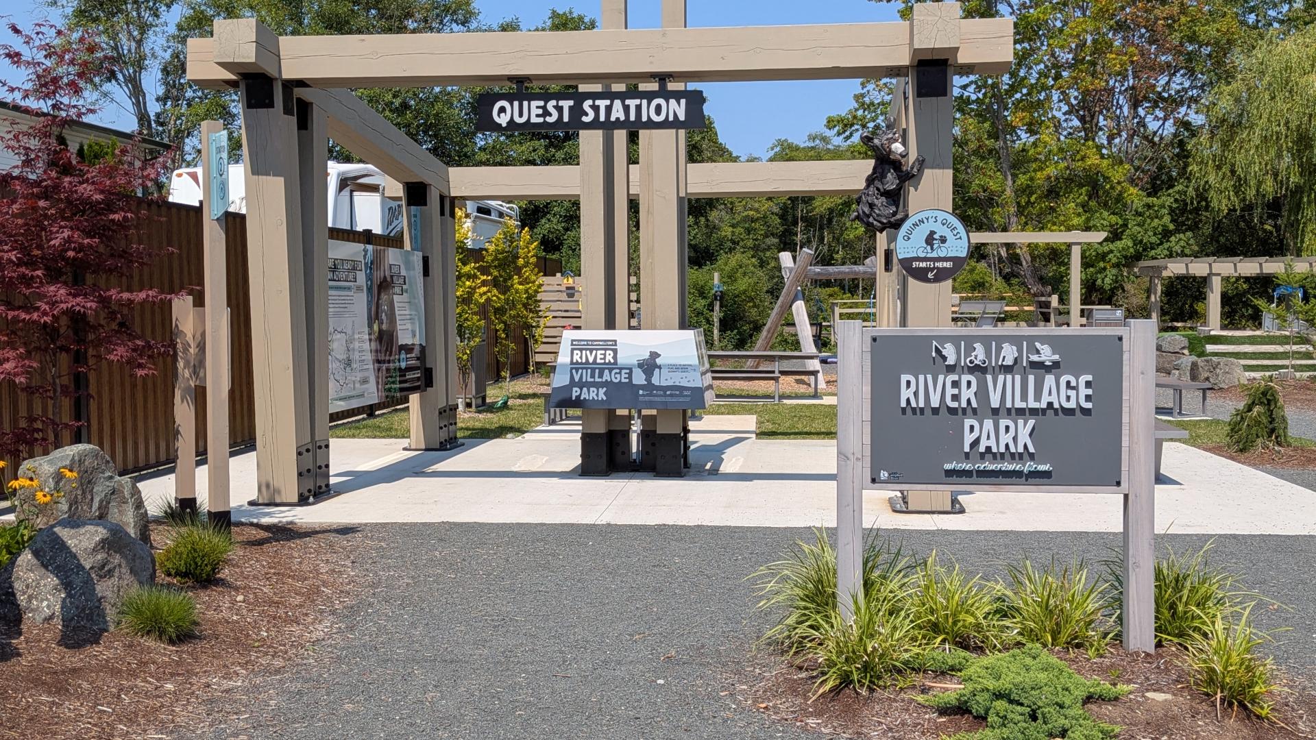 Alt text: Entrance to River Village Park on a sunny day, featuring a wooden pergola with a “Quest Station” sign, interpretive displays, and a large River Village Park sign. Landscaped gardens, trees, carved wooden sculptures, and playground features are visible throughout the park setting.