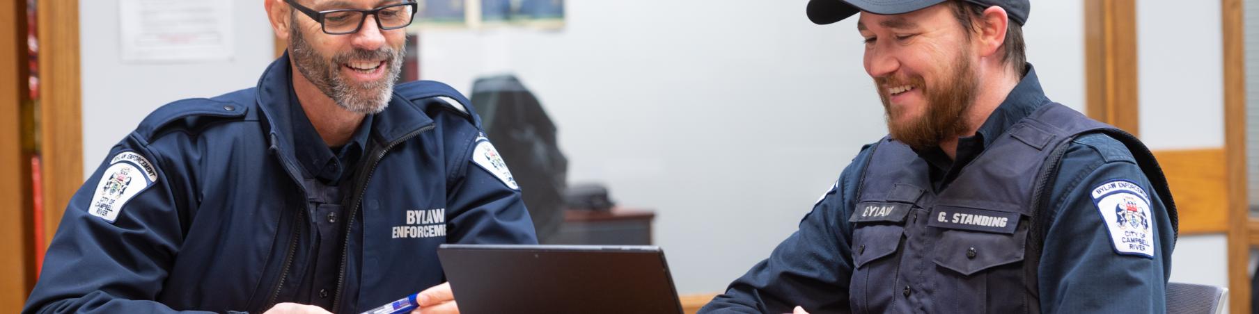 Two uniformed officers smiling at a table, using a tablet.