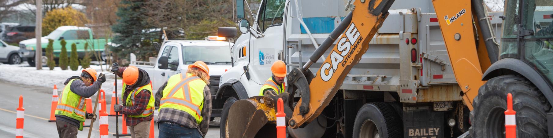 Workers in safety vests repairing a road with a backhoe and traffic cones.