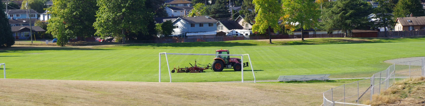 Driving lawn mower mending one of the baseball fields behind the Sportsplex on a sunny day.