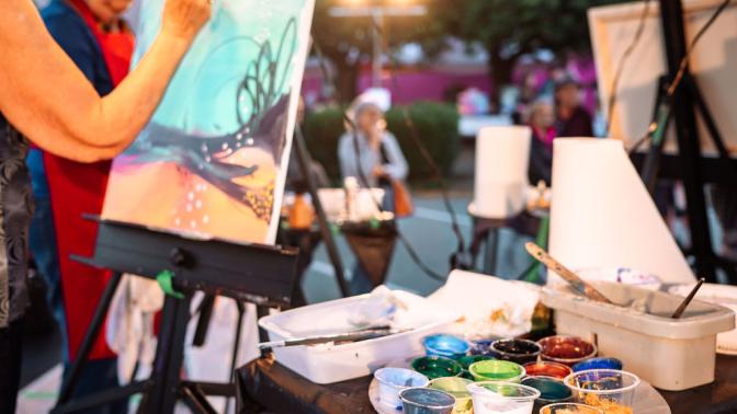 Artist painting on canvas, surrounded by paint jars and brushes, in an outdoor setting.
