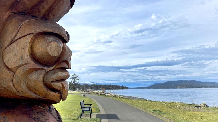 Indigenous carving facing the Discovery Passage with a trail and bench in the background.