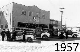 Fire trucks and firefighters outside a brick fire hall, 1957.