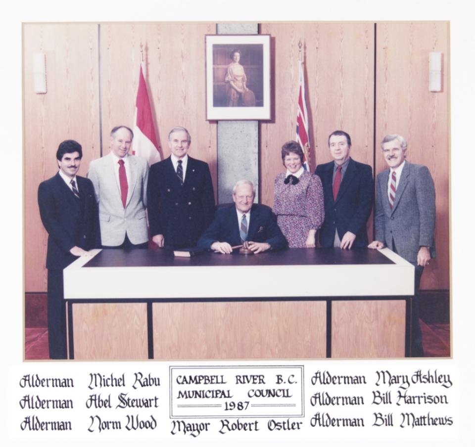 Council members standing around a seated man at a desk, flags in the background.