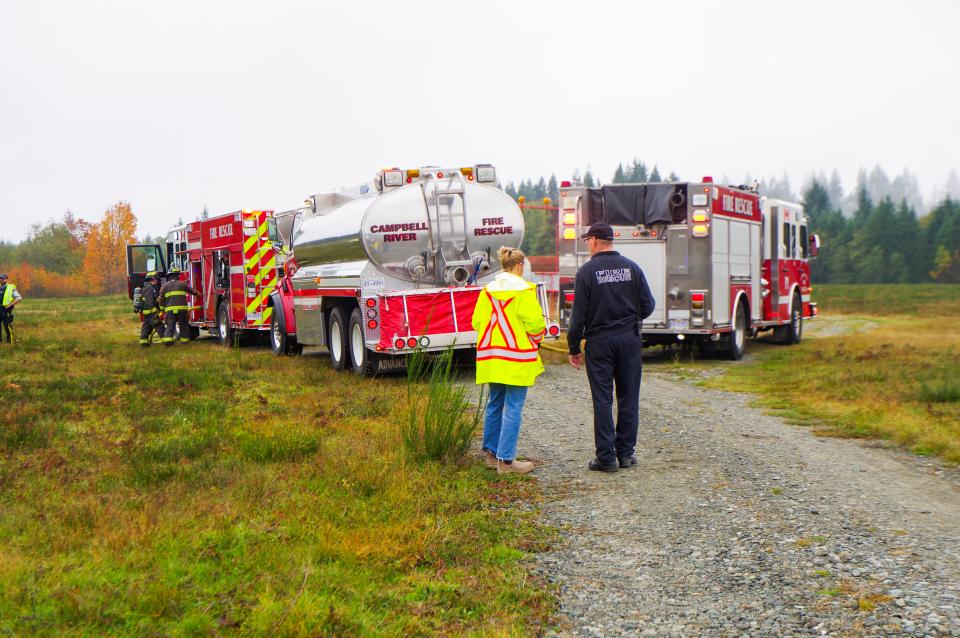 Two fire trucks, a water truck are parked by each other in a field and people in safety gear are having a conversation.
