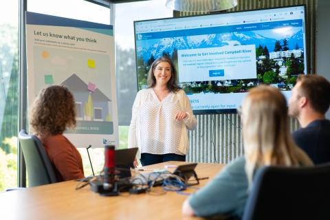 Four people participate in a meeting. One is smiling and addressing the group, leading a presentation. There is a feedback board. The Get Involved Campbell River website is displayed on a monitor. There is a large window letting in bright light.