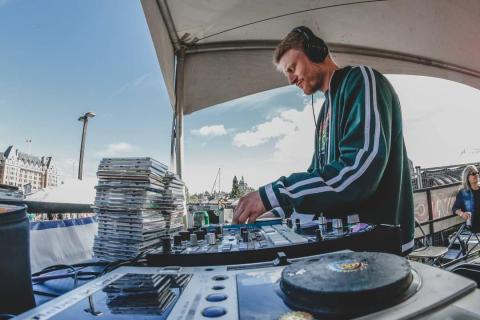 DJ Rebel Selector performs on an outdoor stage. Equipment such as turntables and CDs are onstage for Rebel Selector's use.