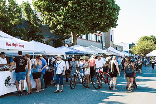 People walking through a street market in Campbell River, surrounded by vendor booths selling local goods and food, with a lively, community atmosphere.