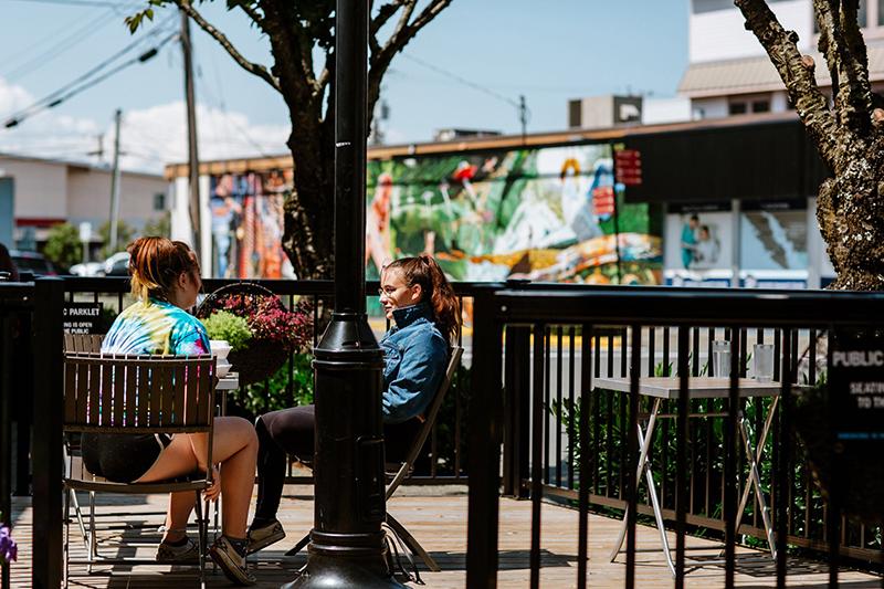 Two people sitting and talking at a parklet patio outside a downtown business.