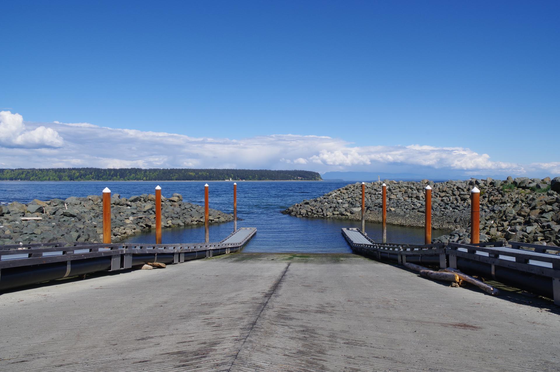 A view looking down the paved boat ramp at Big Rock Boat Ramp in Campbell River. The ramp leads into the ocean. An island is in the background, across the water. It is a sunny day and the sky is blue with some clouds on the horizon.