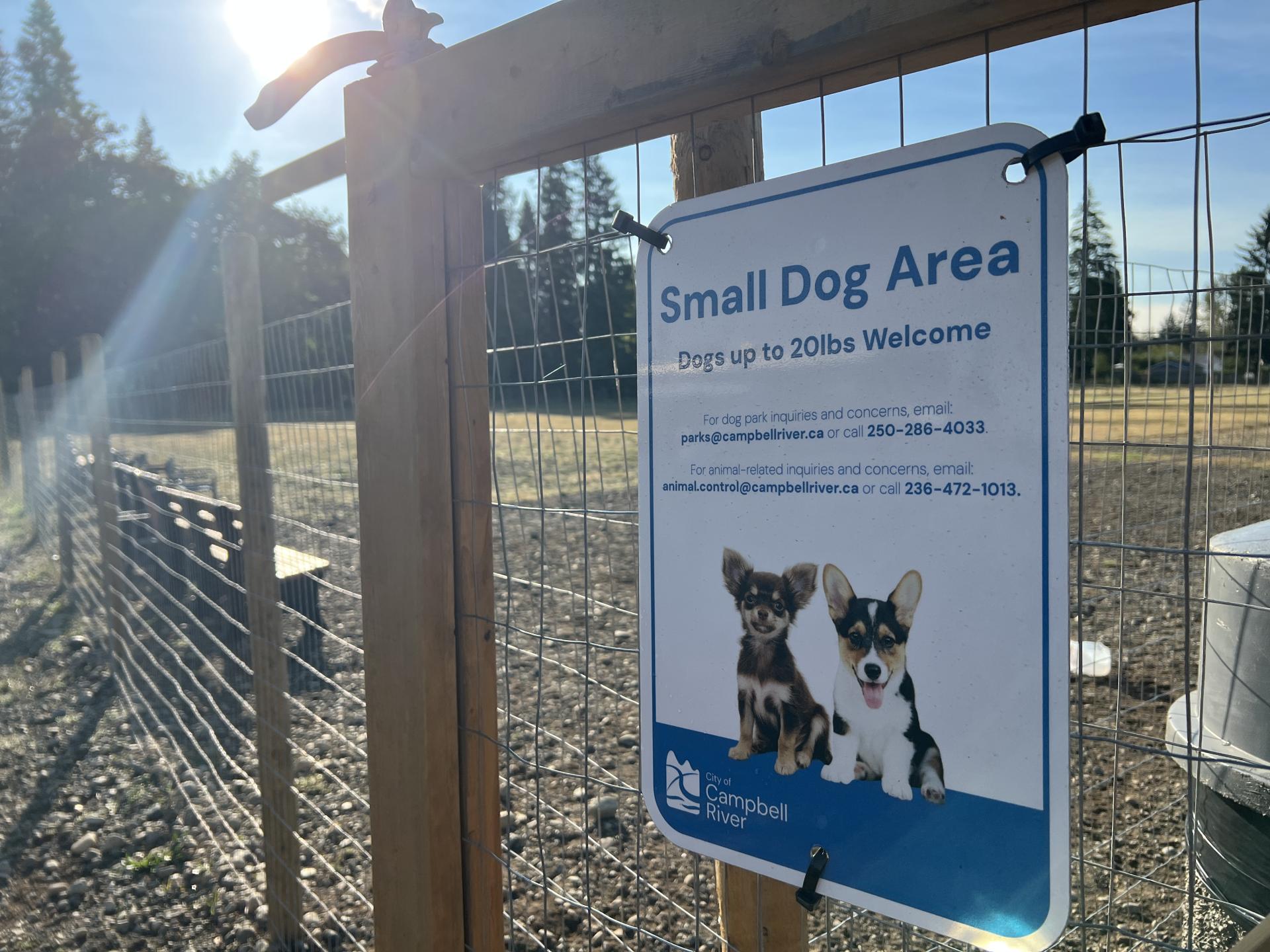 A fenced-in grassy area at Evergreen Dog Park. A sign indicates that this is an area for small dogs. It is a sunny day and the sky is blue. Trees are in the background.
