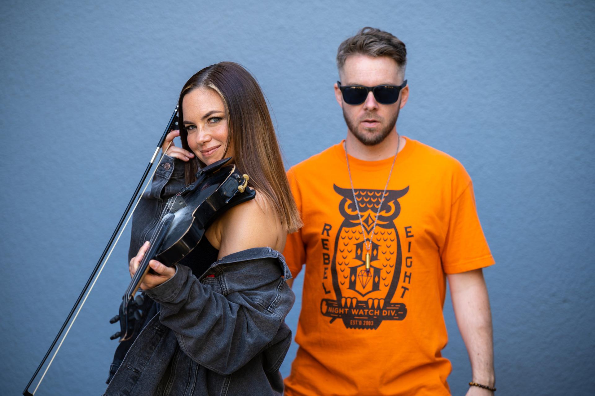 Musicians Kytami and Phonik Ops pose together against a blue background. Kytami is holding a violin. The photo was provided courtesy of Kytami and Phonik Ops.