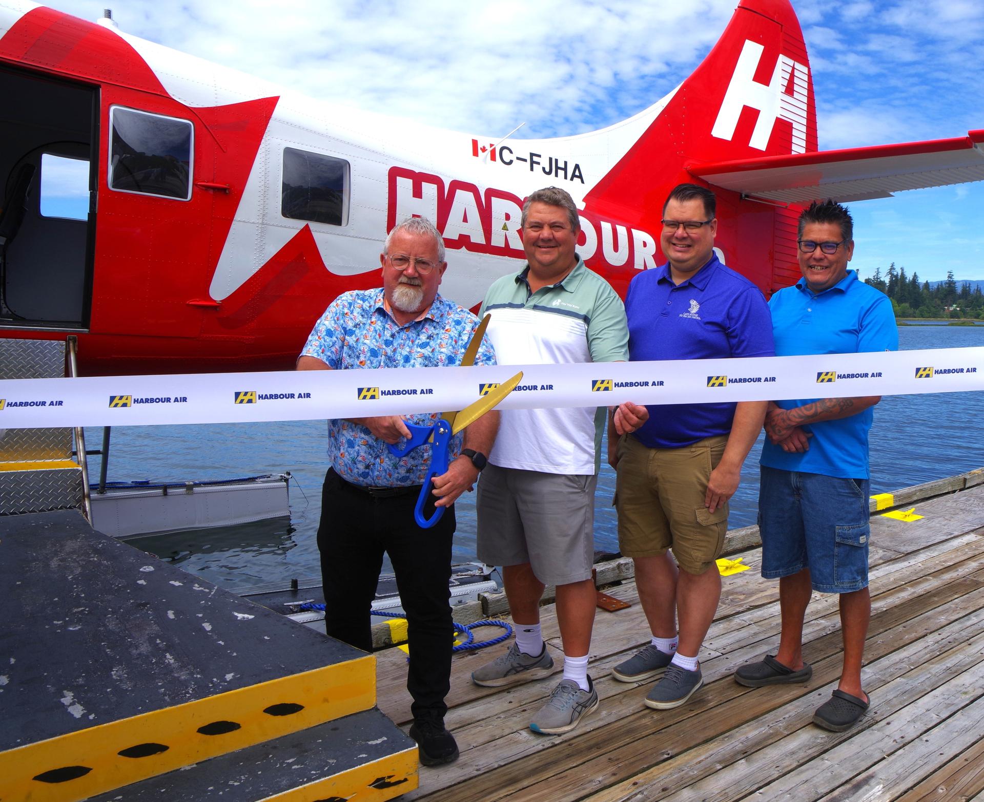 Mayor Kermit Dahl, Chief Chris Roberts, Jason Wilson and Andrew Puglas are gathered behind a ribbon, which Mayor Dahl is about to cut with a giant pair of scissors. A Harbour Air plane is in the water behind them.